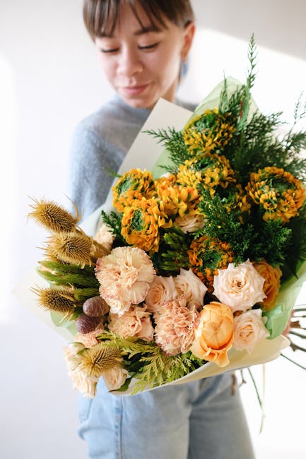 A young person with short brown hair is holding a large bouquet of fresh flowers wrapped in light green paper. The bouquet features a vibrant mix of yellow marigolds, pale pink carnations, soft peach