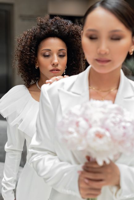 A close-up of a bride in a white dress holding a bouquet of pale pink and white roses, tightly arranged with some greenery, wrapped in light pink paper and tied with a matching ribbon. She stands indo