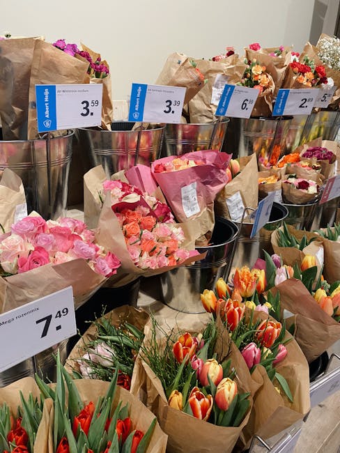 A display of various flower bouquets in a floral shop, arranged in metal buckets and wrapped in brown paper. The bouquet at the front features a mix of red and pink tulips with green stems and leaves,