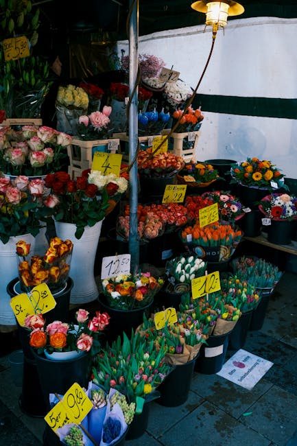 An outdoor display of fresh flower bouquets arranged in black and white plastic buckets on a market stall. The flowers include pink and white roses, orange and yellow tulips, multicolored gerbera dais