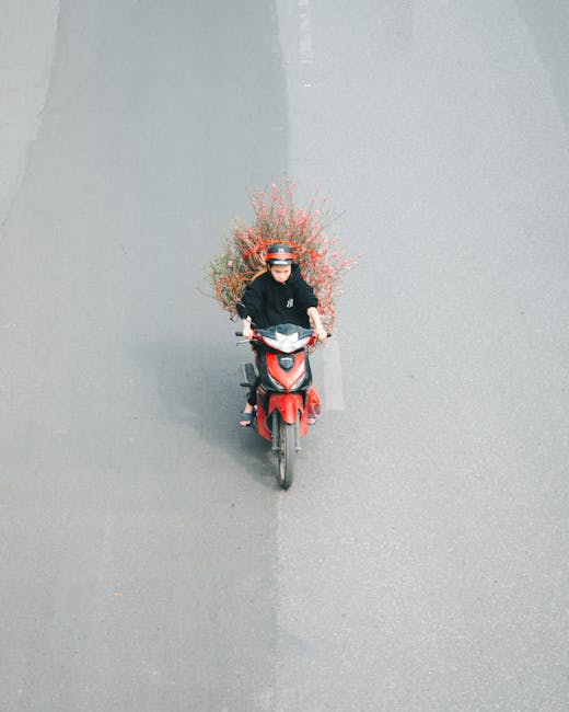 A delivery person riding a red scooter on a paved street, carrying a bouquet arrangement of fresh flowers behind them. The bouquet features vibrant pink, red, and orange blossoms, with green foliage, 