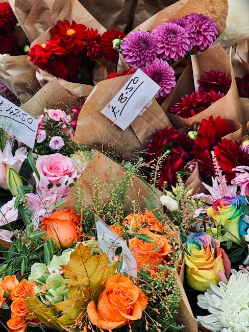 A vibrant display of fresh floral bouquets arranged in brown paper wrapping, featuring a variety of flowers including bright red dahlias, purple chrysanthemums, soft pink roses, orange roses, multicol