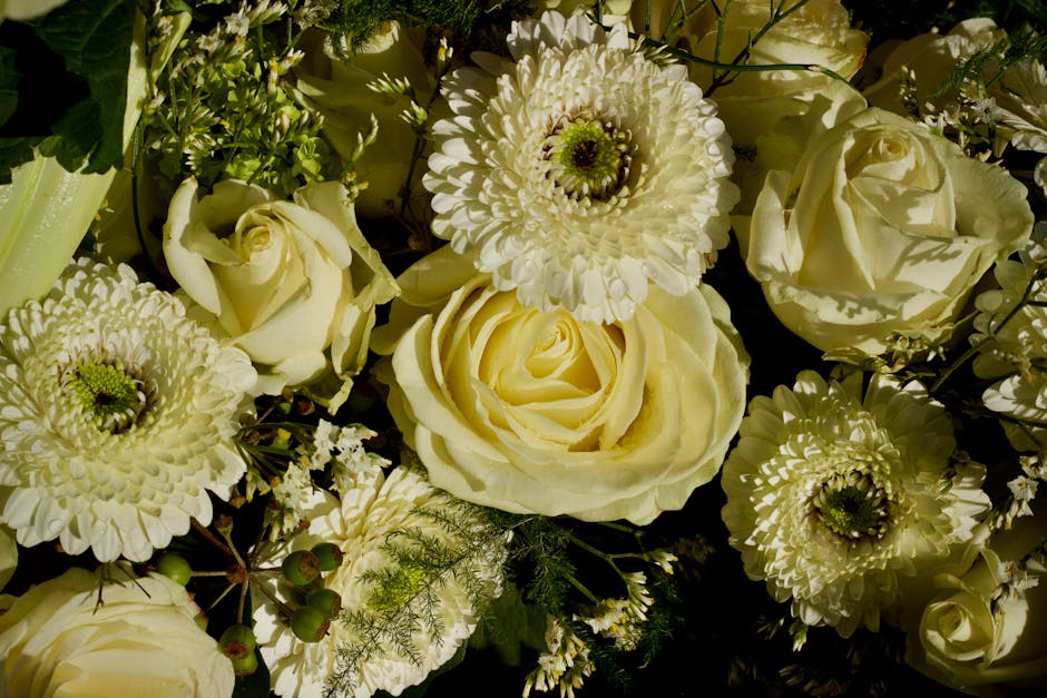 A close-up arrangement of fresh cream and white flowers, including roses, chrysanthemums, and other floral varieties, with detailed petals and green stems visible. The bouquet features a mix of open b