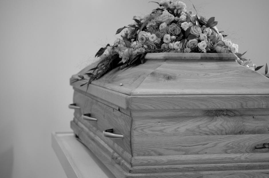 A wooden funeral casket with a smooth, polished surface displaying natural wood grain, adorned with a floral arrangement of mixed roses, lilies, and other blooms in soft pastel shades, including white