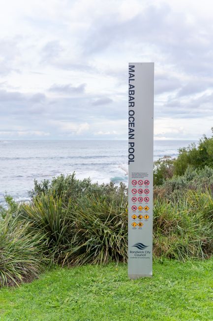 A tall, vertical signpost reading 'Malabar Ocean Pool' stands amidst lush green foliage and tall grasses on a coastal landscape, with the ocean in the background under a cloudy sky. The sign features 